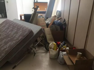 Cluttered room filled with an old mattress, furniture, cardboard boxes, and miscellaneous household junk.